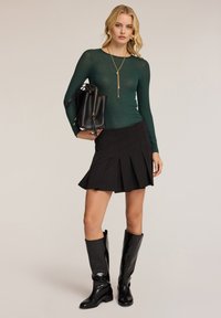 Vihreä pitkähihainen paita, mustat laskostetut hameet, mustat polvipituiset saappaat. Malli pitää mustaa käsilaukkua, jossa on kultainen yksityiskohta hihnassa. Sujuvat tekstuurit.