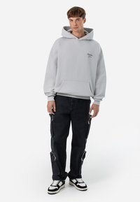 Lys grå hættetrøje med en frontlomme og logo. Sorte cargo bukser med sidelommer med lynlås. Sorte og hvide sneakers fuldender outfittet.