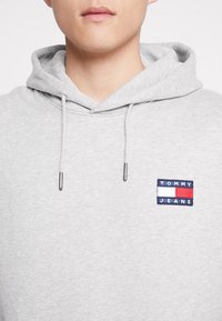 Primo piano di una persona che indossa un felpa grigia chiara con un logo sul davanti che dice "Tommy Jeans" in blu, rosso e bianco.