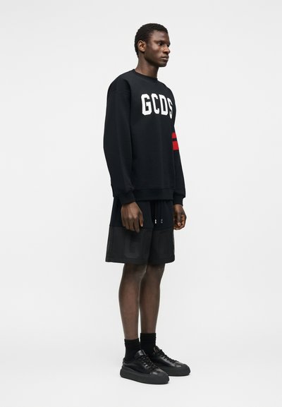 Άνδρας που φοράει μαύρο φούτερ GCDS με κόκκινες ρίγες στο μανίκι, μαύρο σορτς, μαύρες κάλτσες και μαύρα αθλητικά παπούτσια, στέκεται πλαγίως σε λευκό φόντο.