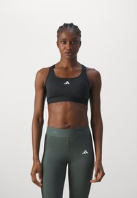 Zwarte sportbeha met wit logo, met een ronde halslijn en brede banden, gecombineerd met donkergroene high-waisted legging met accenten.