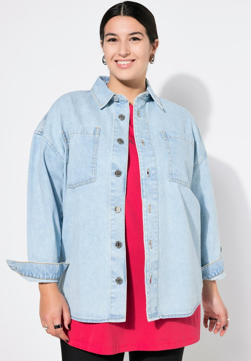 Chemise en denim bleu clair surdimensionnée avec deux poches avant, boutons argentés et manches retroussées, superposée sur un top rouge. Design lisse et décontracté.