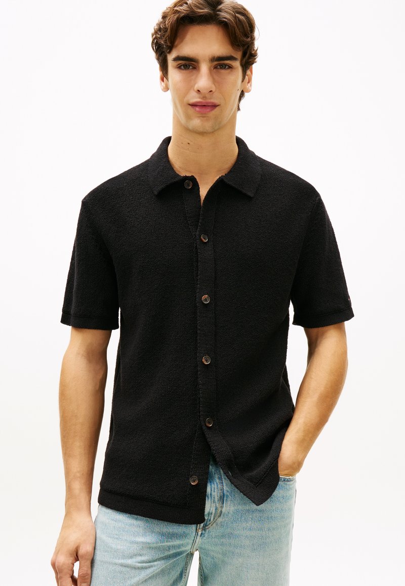 Νέος άνδρας που φοράει μαύρο κοντομάνικο πουκάμισο με κουμπιά και ανοιχτόχρωμο μπλε τζιν, στέκεται με ένα χέρι στην τσέπη του.