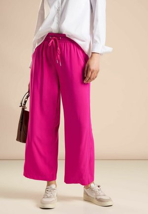 Person wearing bright pink wide-leg pants, white sneakers, and a white button-up shirt holding a dark brown handbag.