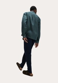 Camisa verde de botones y mangas largas, pantalones azul marino y zapatos marrones sin cordones. La camisa tiene un corte relajado y una textura suave.