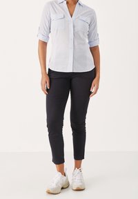 Kvinna i ljusblå skjorta med knappar, svarta slim-fit byxor och vita sneakers, stående mot en helvit bakgrund.