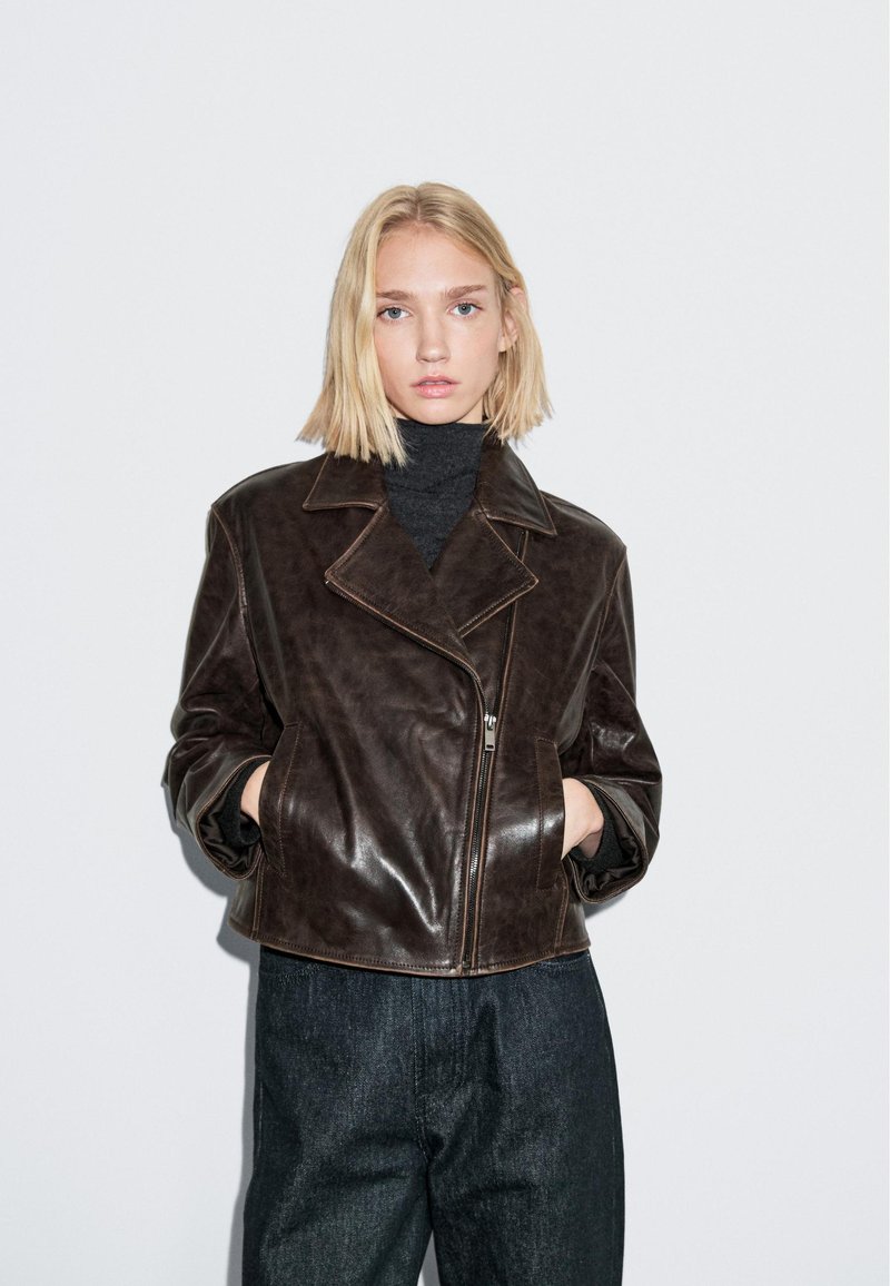 Braune Lederjacke im Cropped-Stil mit großem Kragen, Reißverschluss und Seitentaschen. Kombiniert mit dunkelblauen Jeanshosen und zeigend einen legeren Stil.