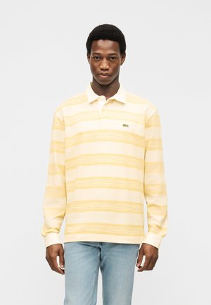 Férfi, aki sárga és krémszínű csíkos hosszú ujjú galléros pólót és világoskék farmert visel, sima fehér háttér előtt áll.