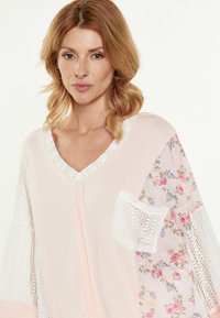 Lichtroze tuniek met bloemenpatroon op de mouwen en een textuurkant zakje. Heeft een V-hals en een losse pasvorm voor comfort.