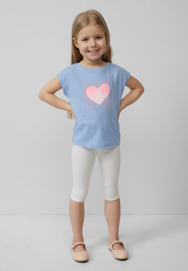 Lichtblauwe t-shirt met een roze hartgrafiek en tekst, gekleed met witte leggings en lichtroze schoenen. Glad, casual stof en pasvorm.