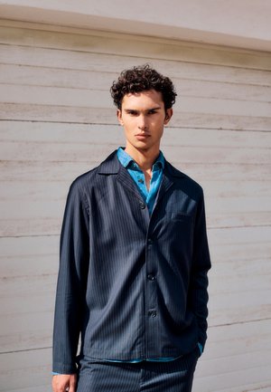 Fiatal, göndör sötét hajú férfi sötét csíkos zakót visel kék ing fölött, világos fa háttér előtt állva.