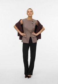 Gilet marron ajusté avec deux poches avant et boutons noirs, superposé sur un châle bordeaux foncé. Associé à un pantalon slim noir et des chaussures pointues.