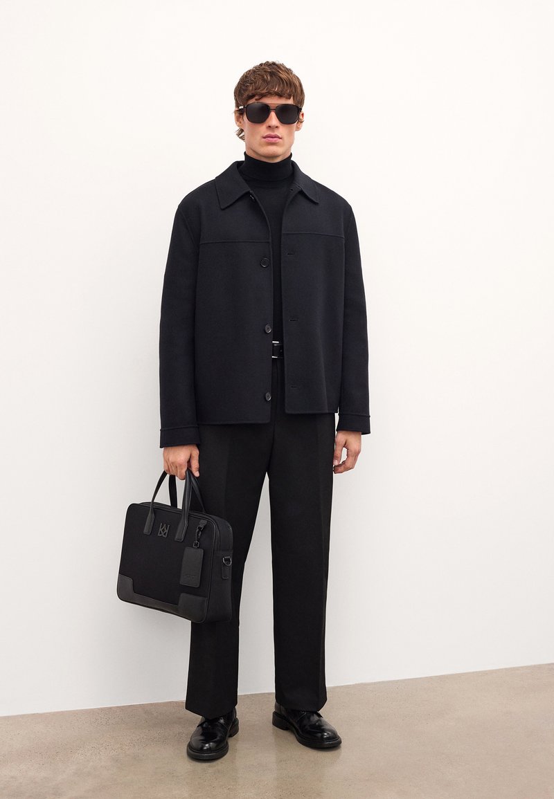 Fekete kabát gallérral egy fekete garbón, széles lábú fekete nadrággal párosítva. Fekete bőr táskát tart, kiegészítőkkel. Fekete napszemüveg.