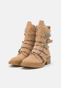 Botas de tornozelo em camurça bege com múltiplas correias, fivelas de metal e um salto bloco. Apresentam uma superfície texturizada e detalhes costurados.