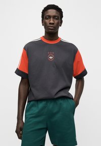 Γκρι και κόκκινο κοντομάνικο μπλουζάκι με στρογγυλή λαιμόκοψη, με κεντρικό λογότυπο και τρία αστέρια από πάνω· συνδυασμένο με πράσινα σορτς.