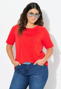 Červené tričko s krátkými rukávy a potiskem "TRUE size" v tmavých písmenech, zkombinované s modrými džíny a světle modrým pásem. Hladká textura.