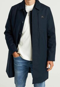 Man i en mörkblå knäppt kappa över en vit skjorta och blå jeans, med en hand i fickan mot en enfärgad bakgrund.