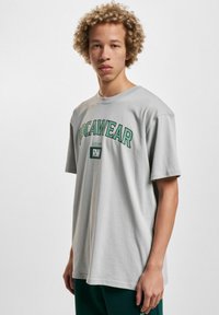 Vaalea harmaa puuvillapaita, jossa on vihreä "ROCAWEAR" -teksti ja logo. Tässä paidassa on lyhyet hihat ja pyöreä kaula-aukko; rento leikkaus.
