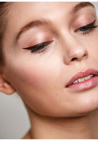 Nahaufnahme eines Gesichts mit heller Haut, leichtem Make-up, zartem Rouge, definierten Augenbrauen und dunklem Eyeliner, der die Augen betont. Die Lippen sind leicht rosa getönt.