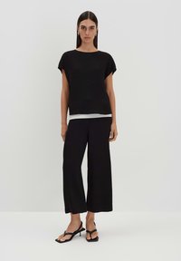 Top nero a maniche corte con vestibilità comoda e scollatura rotonda, indossato sopra una base bianca. Accoppiato con pantaloni neri a gamba larga e sandali neri.