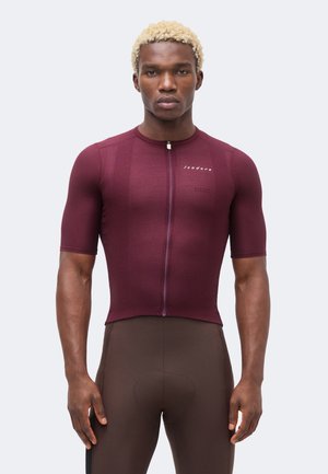 Rövid szőke hajú férfi, bordó, testhez simuló kerékpáros mezben és sötétbarna párnázott kerékpáros rövidnadrágban, sima háttér előtt áll.