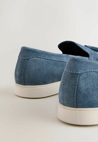 Sinised seemisnahast libisevad mokassiinid valgete kummist talladega, vaadatuna tagantpoolt heledal neutraalsel taustal.