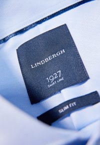 Interno di una camicia azzurra chiaro con etichetta navy su cui sono scritti in bianco "Lindbergh," "1927," "Shirt Line," e "Slim Fit." Texture del tessuto liscia.