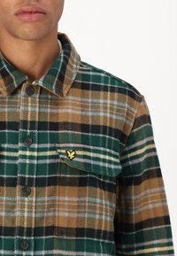 Prugasta košulja u zelenoj, smeđoj i crnoj boji. Flanel materijal s kopčanom produžetkom i džepom na prsima ukrašenim žutim logotipom.