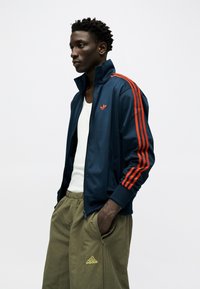 Νέος άνδρας με μπλε τζάκετ με κόκκινες ρίγες και μπεζ παντελόνι Adidas, στέκεται με τα χέρια στις τσέπες μπροστά σε απλό λευκό φόντο.