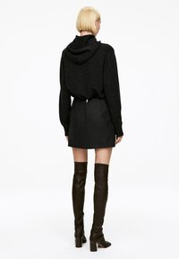 Schwarzer Hoodie mit lockerer Passform und Kapuze, kombiniert mit einem grauen Minirock mit einem Reißverschluss auf der Rückseite. Das Model trägt kniehohe braune Stiefel.