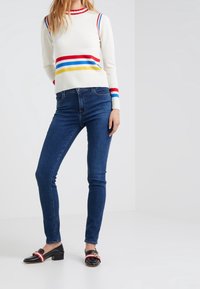 Vit stickad tröja med röda, blå och gula ränder, ihop med blå slim fit jeans och svarta slip-on skor med röda accenter.