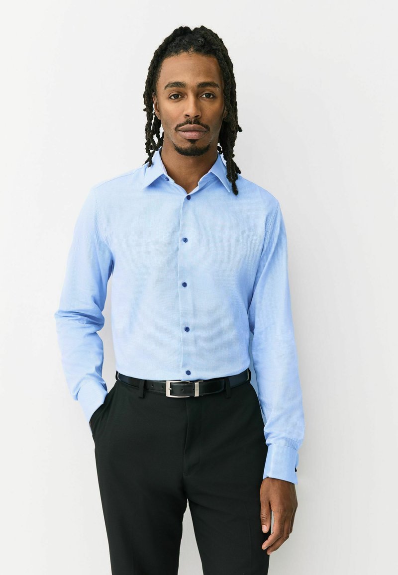 Svetlomodrá košeľa s dlhým rukávom a textúrovaným vzorom, zapnutá na gombíky, s kontrastnými tmavomodrými gombíkmi, v kombinácii s čiernymi nohavicami a opaskom.