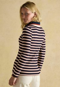 Sieviete ar gaišiem matiem, ģērbusies zili, sarkani un baltā svītrainā garajās piedurknēs topā un gaišos biksēs, stāv pretī bēša sienai.