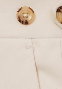 Tissu beige avec deux grands boutons bruns texturés en spirale et des accents de couture visibles. Gros plan sur les détails du bouton et du tissu.
