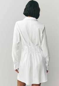 Vit skjortklänning med långa ärmar, figurnära bodice, veckad baksidesdesign och knäppte manschetter; slät tygtextur.