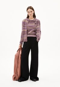 Femme portant un cardigan rayé violet sur un pull assorti, un pantalon large noir, tenant un blazer rose dans sa main gauche, se tenant sur fond blanc.