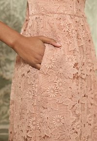 Rosa spetsklänning med blommönster, intrikata detaljer och en sidficka. Tyget är strukturerat och genomskinligt, vilket framhäver mönstret.