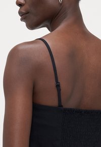 Haut noir sans manches avec bretelles réglables, présentant un panneau arrière froncé et une texture de tissu lisse, mettant en valeur la zone des épaules.