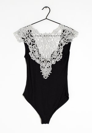 Une combinaison noire avec un col en dentelle blanche complexe, présentant des motifs floraux et des accents de perles. Suspendue à un crochet noir.