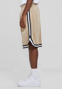 Beige Netzshorts mit schwarzen und weißen Seitenstreifen, die Schlitze am Saum aufweisen. Kombiniert mit weißen Sneakers und Sneakersocken.