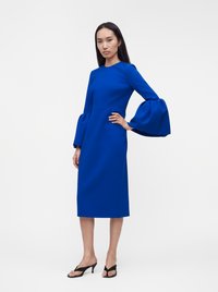 Femme aux longs cheveux noirs portant une robe midi bleue à larges manches cloche et des sandales noires à talons, debout, la main sur la hanche.