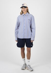 Femme portant une chemise à rayures verticales bleues et blanches boutonnée, un short bleu marine, des chaussettes blanches, des baskets et une casquette de baseball blanche avec un logo.