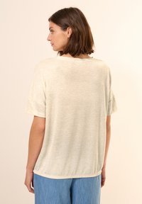 Maglietta corta a maniche, di un beige chiaro, con una vestibilità comoda, texture morbida, scollo rotondo e orlo leggermente curvato. Abbinata a pantaloni blu chiaro.