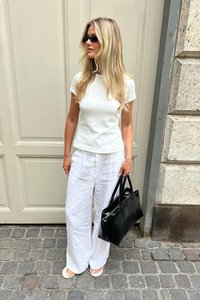 Balta pieguļoša T-kreklu, brīvi baltie lina bikses, melna ādas rokassoma un baltas sandales. Cirtaini mati, saulesbrilles, stāvot pret neitrālu fonu.