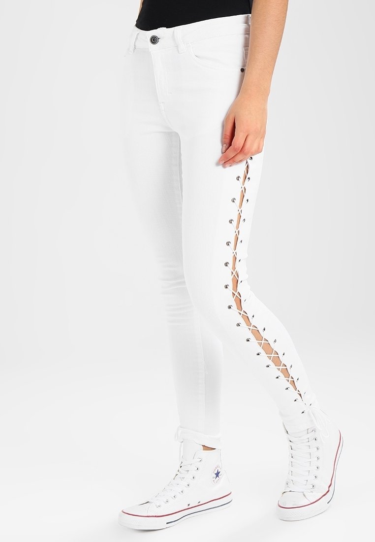 Jeans skinny blancos con cordones laterales y ojales plateados, que presentan un diseño ajustado y dobladillos enrollados, combinados con zapatillas blancas.