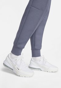 Sneakers bianche Nike con tomaia in mesh e ammortizzazione aerea visibile. Indossate con pantaloni della tuta grigi con taglio affusolato, tasche laterali e polsini a costine.