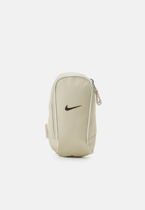 Pochette beige Nike avec logo swoosh noir, compartiment principal zippé et bordure blanche sur un fond blanc uni.
