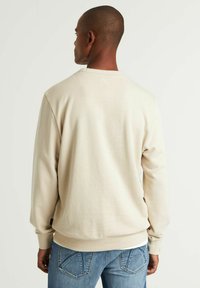 Sudadera beige con un tejido texturizado, puños acanalados y una cinturilla. La parte de atrás presenta un detalle de costura sutil y se combina con jeans azules.
