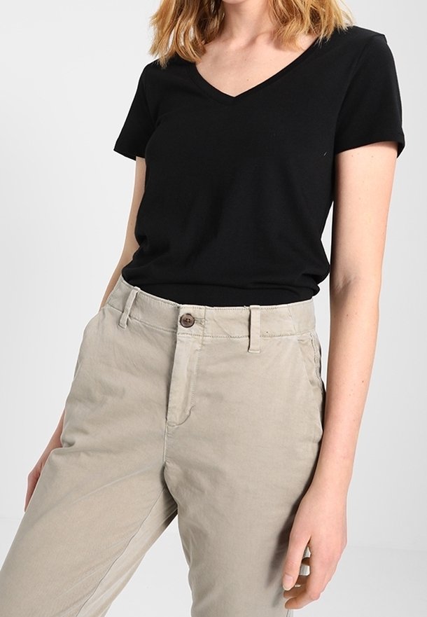 Femme portant un t-shirt noir à manches courtes et col en V, rentré dans un pantalon beige taille haute avec fermeture par bouton et zip.