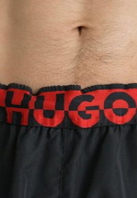 Fekete shorts rojtos piros derékpánttal. A derékpánton a "HUGO" márkaneve vastag fekete betűkkel szerepel élénk piros háttérrel.